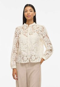 Blouse en dentelle blanc cassé à manches longues, motif floral, col montant et fermeture à boutons sur le devant. Tissu léger et semi-transparent.