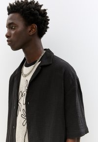 Camicia nera strutturata con colletto indossata sopra una t-shirt bianca con motivo. Sembra sovrapposta con una collana, enfatizzando un taglio rilassato.