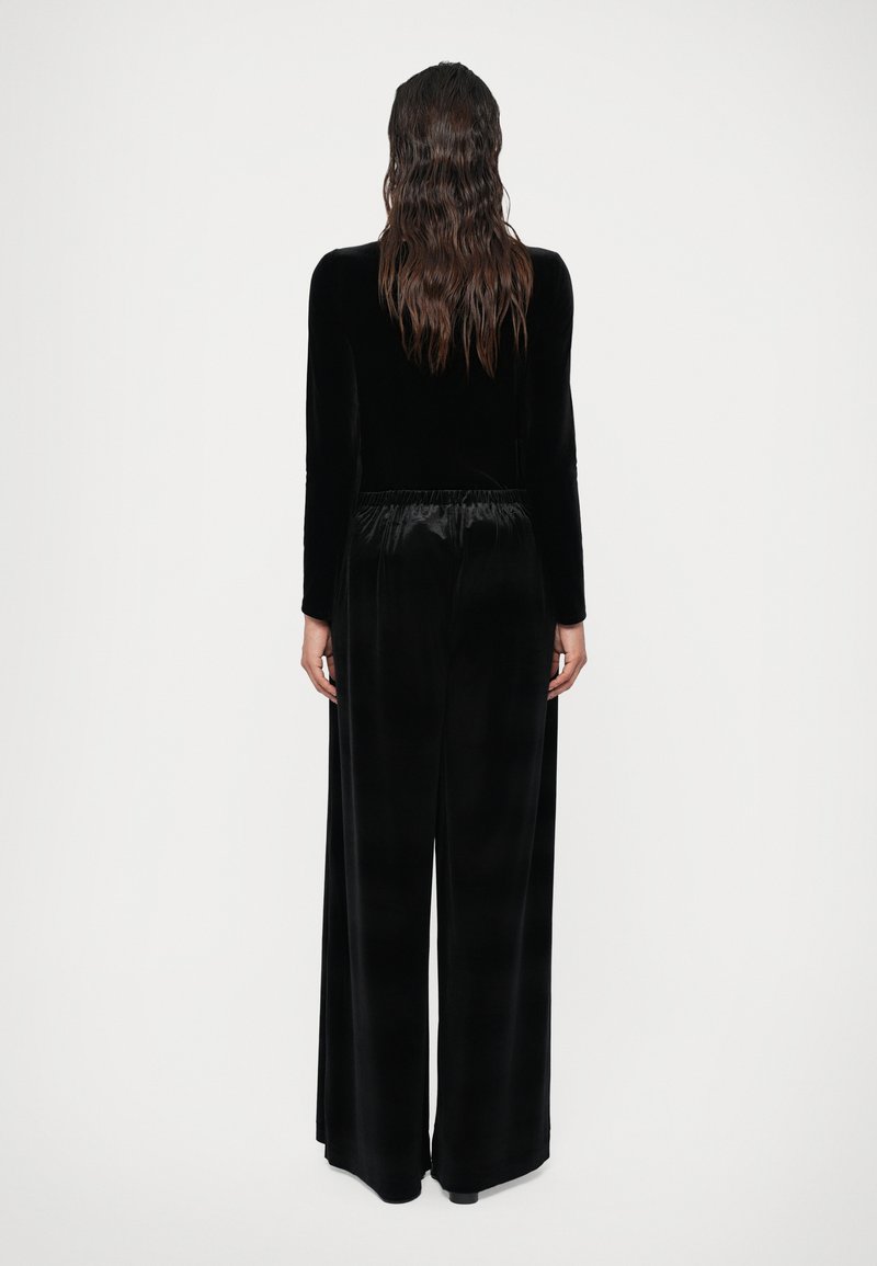 Black velvet jumpsuit with long sleeves, wide-leg design, and a cinched waistband. Smooth texture with no visible patterns or embellishments.