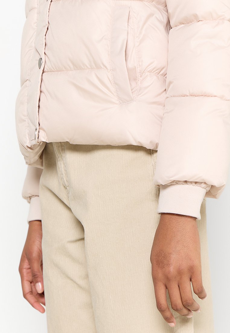 Lichtroze pufferjack met een cropped ontwerp, met een voorzak en ribgebreide mouwmanchetten. Draagt over beige corduroy broek.