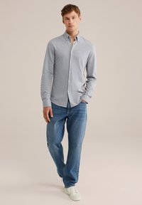 Hombre caminando vestido con una camisa de botones gris claro, jeans azules y zapatillas blancas frente a un fondo beige.