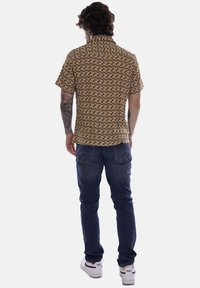 Camicia a maniche corte con colletto marrone con un motivo circolare, abbinata a jeans blu e sneakers bianche. Tatuaggi visibili sul braccio.