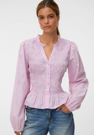 Femme aux cheveux longs porte un chemisier rayé rose et blanc boutonné avec des manches longues bouffantes et un jean bleu, main dans la poche.