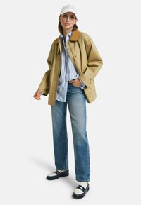 Giacca beige con colletto marrone, camicia a righe blu, jeans azzurri chiari e mocassini neri. Un cappellino bianco completa il look.
