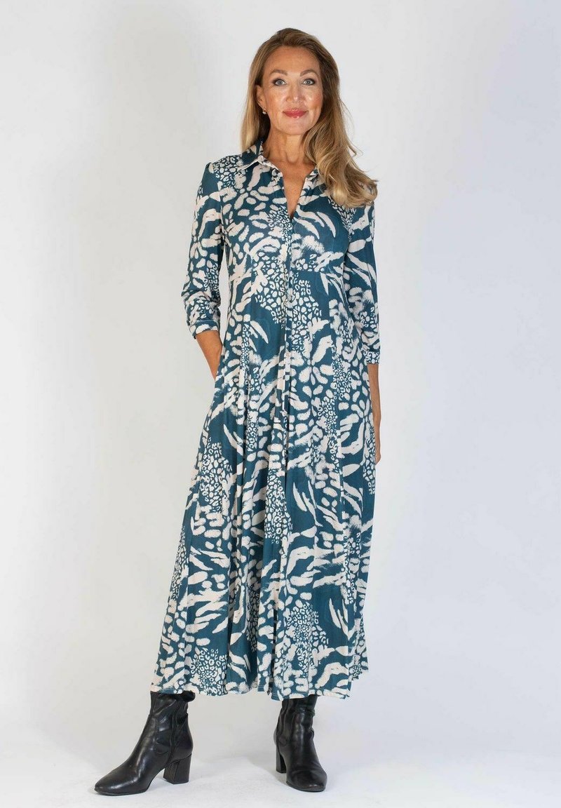 Długa, zwiewna sukienka w odcieniu teal z białymi organicznymi wzorami, z kołnierzykiem i długimi rękawami; połączona z czarnymi botkami za kostkę.