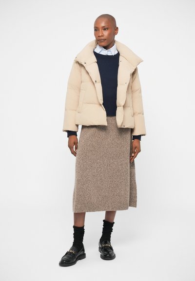 Beige puffer jacket with a fold-over collar, navy knitted sweater underneath, and a long, textured brown skirt. Black loafers with a gold accent.