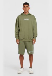Oliven grøn hoodie med en kængurulomme, afslappet pasform og logo tekst. Parret med matchende shorts med sidelommer og logo. Hvide sko.