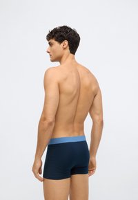Young man standing shirtless facing left, wearing dark blue boxer briefs with a light blue waistband against a plain white background.