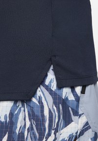 Gros plan de l’ourlet d’un vêtement bleu marine avec une petite fente latérale, superposé à un tissu à motifs abstraits bleus et blancs en dessous.