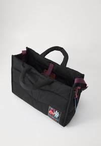 Svart polstret tote-veske med lilla hender, med et fargerikt brodert merke og indre lommer. Teksturert stoff, rektangulær form.