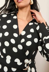 Schwarzes Wickelkleid mit großen weißen Punkten, ausgestattet mit einem Kragen und einer taillierten Taille mit einem metallischen Akzent. Glatte Stofftextur.