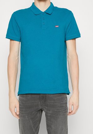Mann trägt ein türkisfarbenes kurzärmeliges Polohemd mit Kragen und rotem Levi's-Logo auf der Brust, kombiniert mit dunkelgrauen Jeans.