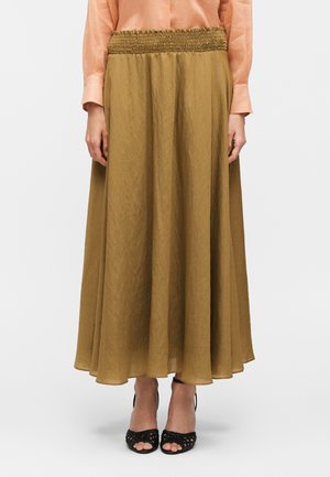 Femme portant une longue jupe fluide couleur moutarde avec une taille froncée, associée à un chemisier pêche et des talons ouverts noirs.