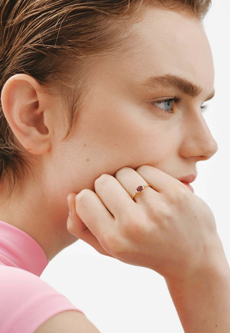 Anillo de oro con una gema roja central, banda texturizada, llevado en una mano que descansa sobre una mejilla. La modelo lleva una blusa rosa y tiene el cabello corto.
