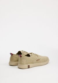 Zapatillas de lona beige con cordones, punta redonda, superficie texturizada y acentos contrastantes en marrón y rojo en el talón y la etiqueta lateral.