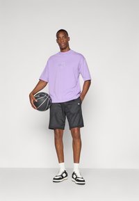 Purple cotton t-shirt with Lakers logo, black mesh shorts, and black-and-white sneakers. Model holds a basketball, standing against a plain background.