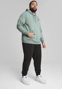 Sweatshirt à capuche vert menthe avec une poche avant, associé à un jogging noir et des chaussures de sport blanches. Présente un logo discret sur la poitrine.
