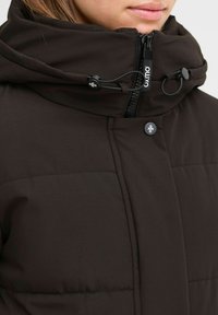 Hnedá prešívaná bunda s vysokým golierom, ktorá má predný zips, nastaviteľné šnúrky a gombíkové akcenty. Štruktúrovaná tkanina, elegantný dizajn.