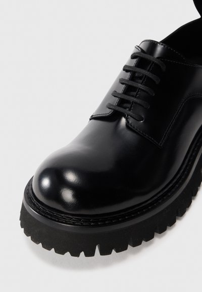 Black leather lace-up shoe with a glossy finish, thick textured sole, and round toe. Features a traditional design with visible stitching.