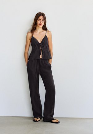 Femeie stând lângă un perete alb, purtând un top negru fără mâneci cu fundă în față, pantaloni negri largi și papuci negri, mâinile în buzunare.