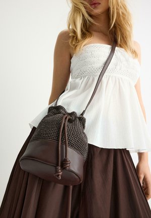 Ženska nosi bel top brez naramnic in rjavo krilo, nosi temno rjavo teksturirano torbico v obliki vedra z usnjenimi poudarki in zavezovanjem z vrvico.