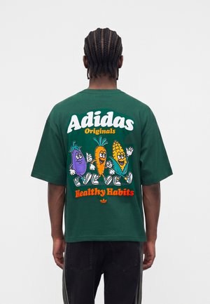 Fonott hajú férfi sötétzöld Adidas pólóban, amelyen rajzfilmes padlizsán, sárgarépa és kukorica látható, sétál, szöveg: „Adidas Originals Healthy Habits.”
