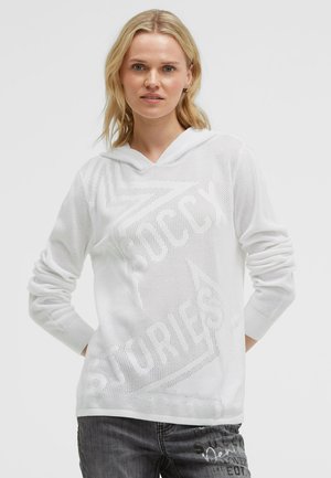 Blonde woman wearing a white mesh hoodie with "Soccy Stories" text and dark gray jeans with printed text, standing against a plain background.