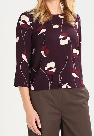 Femme portant un haut violet foncé avec des motifs floraux blancs et bordeaux et des manches trois-quarts, associé à un pantalon marron.
