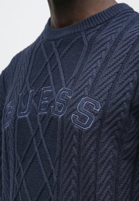 Nahaufnahme eines marineblauen, grob gestrickten Pullovers mit "GUESS"-Stickerei in gleicher blauer Farbe quer über der Brust.