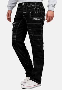 Pantaloni cargo neri con cuciture bianche, multiple tasche, dettagli con zip e vestibilità affusolata. Indossati con scarpe da ginnastica bianche.