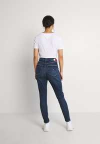 Jeans de tiro alto en azul oscuro con un ajuste ceñido, que cuentan con dos bolsillos traseros y una etiqueta de marca en la cintura, combinados con una camiseta blanca.