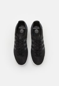 Zapatillas deportivas negras con tres franjas grises, parte superior de tejido texturizado, suela blanca salpicada y logotipo negro redondo en la plantilla.
