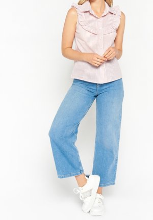 Vrouw die een mouwloze roze-wit gestreepte blouse met ruches draagt, een korte lichtblauwe spijkerbroek en witte sneakers met zilveren accenten.