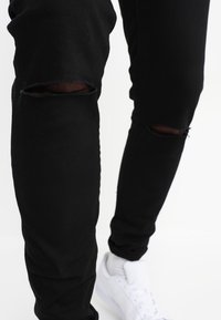 Svarta denim jeans med smal passform och slitna knän, med trasiga partier. Matchade med vita sneakers mot en neutral bakgrund.