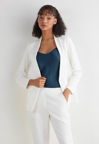Witte op maat gemaakte blazer met lange mouwen, gecombineerd met een donkerblauw satijnen topje. De blazer heeft een reverskraag en een slanke pasvorm.