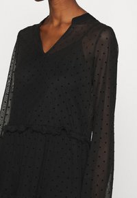Schwarzes, transparentes Kleid mit Punkten, V-Ausschnitt, langen Ärmeln, rüschiertem Taillenbereich und strukturiertem Stoff.