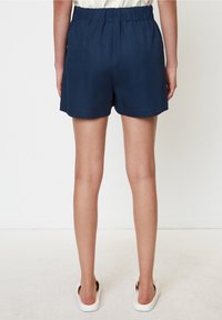 Námořnické modré šortky s elastickým pasem, hladkým povrchem a uvolněným střihem; lem těsně nad kolenem. Nosené s bílými slip-on botami.