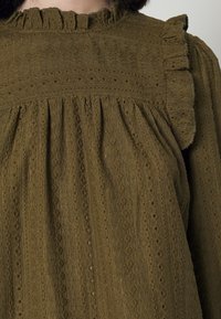Olivgrüne Bluse aus strukturiertem Stoff, mit einem Volant-Halsauschnitt und gefalteten Details, akzentuiert durch kleine, gemusterte Reihen.
