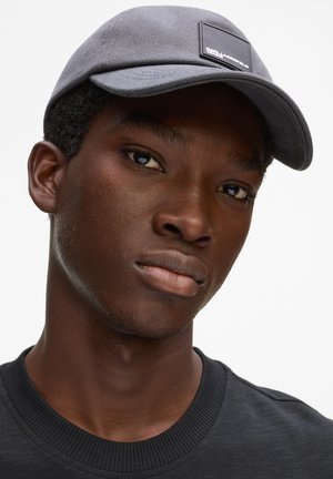 Jauns vīrietis ar tumšu ādu, valkā tumši pelēku cepuri un melnu apaļas apkakles kreklu, skatās nedaudz garām kamerai uz baltas fona.