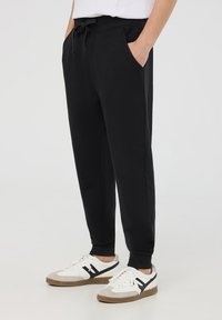 Pantaloni sportivi neri con tasche laterali, vita elastica e cordino. Indossati con sneakers bianche con accenti neri e suola beige.