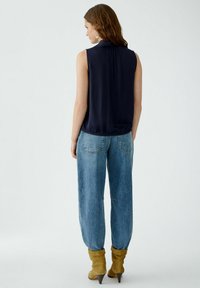 Marineblaue ärmellose Bluse mit Rückfalte, kombiniert mit hellblauen weit geschnittenen Jeans und taupefarbenen, hohen Stiefeletten. Glattes Material und lockere Passform.