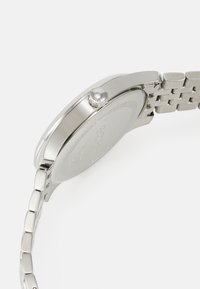 Montre en acier inoxydable avec un boîtier poli, de forme ronde, bracelet en maille métallique, et une couronne proéminente sur le côté pour régler l'heure.