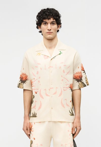 Νεαρός με κυματιστά μαλλιά φοράει μπεζ πουκάμισο με κοντά μανίκια και κουμπιά, καθώς και αντίστοιχα σορτς με ροζ και καφέ αφηρημένα λουλουδένια σχέδια.