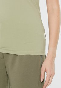 Olivgrünes, tailliertes Tanktop aus glattem Stoff, mit einem seitlichen Etikett "SENSES" und kontrastierenden Nähten am Saum.