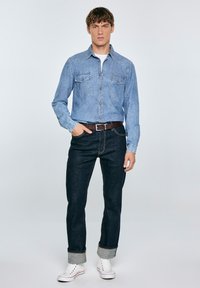 Camisa de mezclilla azul claro con dos bolsillos en el pecho, combinada con jeans azul oscuro enrollados en los puños y zapatillas blancas con acentos rojos.