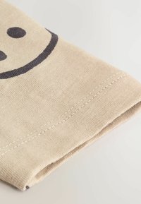 Beige tekstilstof med en synlig søm, præget af et mørkegrå smiley-ansigt design og en glat overflade.