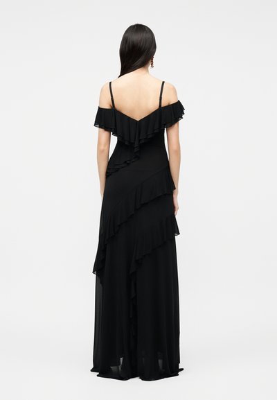 Γυναίκα που φοράει ένα μακρύ μαύρο φόρεμα χωρίς τιράντες με στρώσεις βολάν, όρθια με την πλάτη στραμμένη προς ένα απλό φωτεινό φόντο.