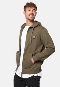 Haut à capuche zippé vert olive en tissu doux, avec un patch avant, deux poches latérales et un cordon de serrage réglable. Porté sur une chemise blanche.
