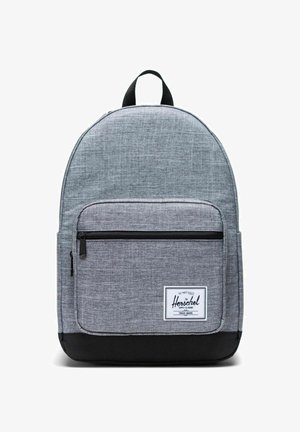 Sac à dos en tissu gris à forme arrondie, poche avant zippée, base noire et étiquette blanche avec le nom de la marque. Matière texturée pour plus de durabilité.