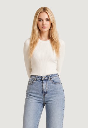 Jonge vrouw met lang blond haar, draagt een witte geribde trui en lichtblauwe hoogwaardige spijkerbroek, staande tegen een effen achtergrond.